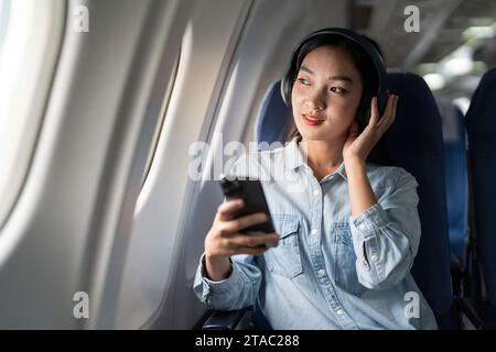 Giovane donna che ascolta la canzone durante il volo in cabina di prima classe utilizzando lo smartphone, la donna si diverte a bordo dell'aereo e ascolta la musica con le cuffie Foto Stock