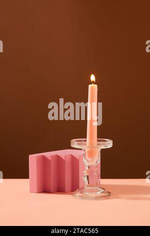 La vista frontale di una candela rosa in fiamme è collocata su un candelabro di vetro e su un podio rosa su un tavolo rosa con sfondo marrone. Spazio per la distribuzione del prodotto Foto Stock