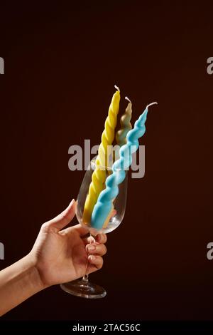 Una mano che regge un bicchiere di vino pieno di candele colorate su uno sfondo marrone profondo. Le candele possono essere fatte con cera d'api, soia, alcune cere vegetali, Foto Stock