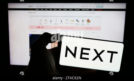 Persona in possesso di un cellulare con il logo della società di vendita al dettaglio di abbigliamento britannica Next plc davanti alla pagina Web aziendale. Concentrarsi sul display del telefono. Foto Stock