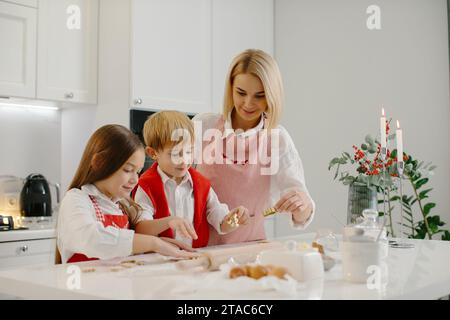 Mamma felice con i bambini che tagliano biscotti di diverse forme in cucina. Madre con figlia e figlio stanno preparando biscotti di Natale a casa e.. Foto Stock