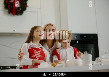 Due bambini felici abbracciano la madre mentre preparano biscotti di Natale a casa durante le vacanze invernali. Madre felice che si diverte con i suoi figli nel ki Foto Stock