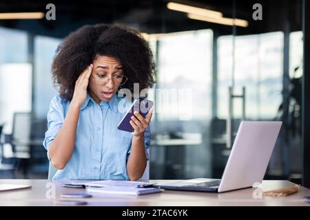 Sconvolta, frustrata e triste donna d'affari, ha ricevuto cattive notizie online al telefono, una dipendente che legge notizie nervosamente, lavora in ufficio con il portatile, donna con i capelli ricci depressi. Foto Stock