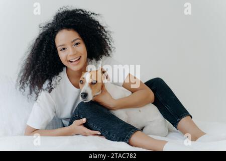 Joyful Black Woman coccola il suo cane su un letto bianco, condividendo un momento felice Foto Stock