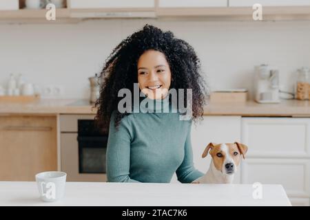 Donna sorridente in dolcevita con il suo cane in una cucina accogliente, una tazza sul tavolo. Foto Stock