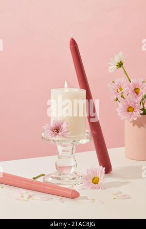 La vista frontale della candela in fiamme è posizionata su un portacandele di vetro. L'area circostante e' decorata con candele colorate e fiori freschi su un bianco e rosa Foto Stock