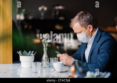 Uomo d'affari vestito con uno smartphone al bar con la maschera inserita, mantenendo la distanza sociale Foto Stock