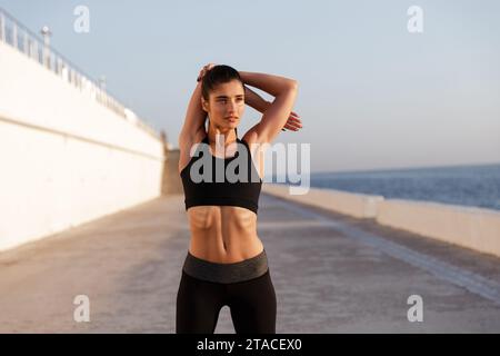 Grave giovane donna sano stretching mani mentre la formazione in mattina vicino al mare Foto Stock