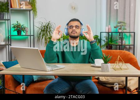 Meditazione, relax, pausa. L'uomo indiano che ascolta la musica respira profondamente, gli occhi chiusi meditano con pensieri concentrati, mente pacifica. Il tizio arabo siede a casa ufficio tavolo posto di lavoro Foto Stock