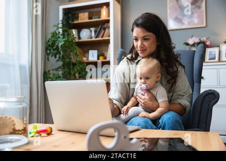 Resta a casa mamma che lavora in remoto sul notebook mentre si prende cura del suo bambino. Una giovane madre d'affari in congedo di maternità che cerca di diventare freelance vicino alla scrivania Foto Stock