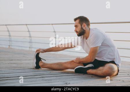 L'intera lunghezza di un giovane uomo atletico dalla barba forte con una t-shirt bianca che si occupa di posare mentre si fa esercizio di stretching per le gambe sedersi a terra a guardare Foto Stock