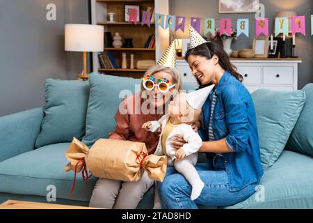 Famiglia multigenerazionale con regali per una festa di compleanno al coperto. Figlia con bambino che festeggia il compleanno di madre o nonna con simpatici cappelli Foto Stock