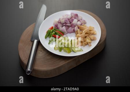 Erbe e verdure a fette per cucinare su un piatto bianco posto su un tagliere di legno. Foto Stock