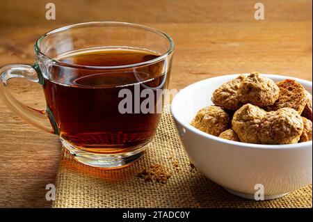 Croccanti e deliziosi biscotti fatti in casa alla meringa di cocco in una ciotola in ceramica bianca con una tazza di tè in vetro trasparente su un panno di legno. Foto Stock