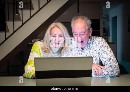 Catturata in un ambiente interno accogliente, una coppia più anziana condivide un momento di piacevole sorpresa e connessione sullo schermo di un notebook. I sorrisi luminosi e gli occhi ampi suggeriscono di essere impegnati in un'esperienza piacevole, magari una videochiamata con i propri cari o scoprendo qualcosa di divertente online. Il calore della camera e il loro abbigliamento confortevole si aggiungono all'atmosfera intima e felice della scena. Collegamento digitale Joyful. Foto di alta qualità Foto Stock