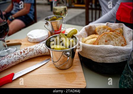 Piccoli sottaceti verdi su salumi rustici e formaggi sparsi su un asse di legno, con i caratteristici mini coltelli rossi svizzeri. Foto Stock