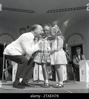 Anni '1960, storico, su una tribuna un'intrattenitrice sul mare con giacca bianca e cravatta di fiocco e tenendo in mano un microfono dell'epoca con due dolci ragazze con sorrisi incantevoli sul palco che chiedono loro di dire qualcosa al pubblico, Inghilterra, Regno Unito. Lettere EH sul palco. Foto Stock