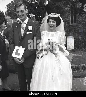 Anni '1960, storico, matrimonio, grandi sorrisi da una coppia appena sposata, fuori dalla chiesa, con la sposa che tiene il ferro di cavallo fortunato e lo sposo con un divertente piatto 'l'' attaccato alla giacca del suo vestito, Inghilterra, Regno Unito. Foto Stock