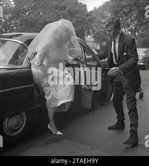 Anni '1960, storico, un giovane ospite di nozze maschile, che indossa una cravatta magra e un winklepickers o "stivali da Beatle", popolare in quest'epoca, tiene aperta la porta dell'auto da sposa per la sposa mentre esce con il suo abito da sposa e velo, Inghilterra, Regno Unito. Foto Stock