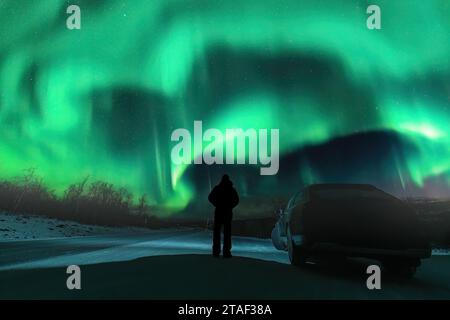 Silhouette di un uomo irriconoscibile e di un'auto scura su una strada di montagna di fronte alle luci verdi del nord che brillano sulle montagne in Svezia, Lapponia. Foto notturna, Foto Stock