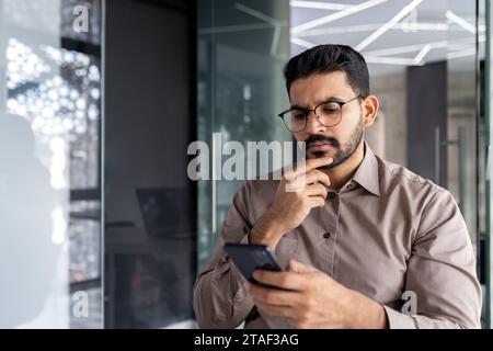 Uomo d'affari che pensa mentre è seduto in ufficio a un tavolo, l'uomo si sta concentrando e leggendo seriamente online utilizzando un'applicazione telefonica, il giovane capo sta tenendo uno smartphone tra le mani. Foto Stock