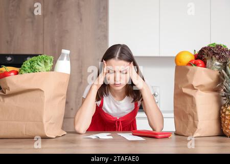 Donna giovane stressata con ricevute del negozio e sacchetti di cibo in cucina. Concetto di aumento dei prezzi Foto Stock