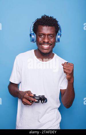 Ritratto di un appassionato giocatore nero maschio con cuffie wireless, sorridere e giocare ai videogame su console. Gioioso uomo afro-americano in abbigliamento casual, che celebra la vittoria tenendo in mano un controller. Foto Stock