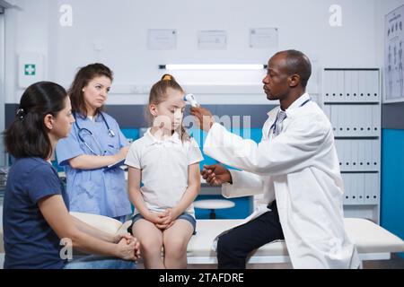 Al check-up medico, il medico usa il termometro per cercare i sintomi del coronavirus. La temperatura della giovane ragazza è misurata da professionisti della salute, che forniscono anche consigli e supporto durante le pandemie. Foto Stock