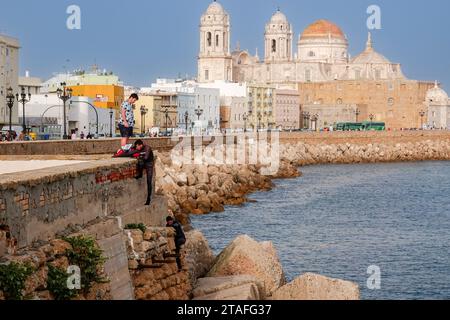 I subacquei si arrampicano lungo l'argine con le cupole della cattedrale di Cadice e la chiesa barocca di Santiago che si innalza sopra il lungomare di Vendaval a Cadice, in Spagna. Foto Stock