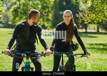 Una coppia beata, adornata con attrezzatura da ciclismo professionale, si gode una romantica passeggiata in bicicletta attraverso un parco circondato da moderne attrazioni naturali Foto Stock