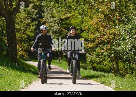 Una coppia beata, adornata con attrezzatura da ciclismo professionale, si gode una romantica passeggiata in bicicletta attraverso un parco circondato da moderne attrazioni naturali Foto Stock