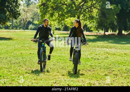 Una coppia beata, adornata con attrezzatura da ciclismo professionale, si gode una romantica passeggiata in bicicletta attraverso un parco circondato da moderne attrazioni naturali Foto Stock