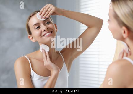 Donna sorridente che si toglie il trucco con tamponi di cotone davanti allo specchio in interni Foto Stock