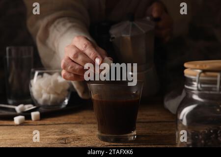 Preparazione di caffè aromatico nella moka. Donna che mette cubetti di zucchero in bicchiere con bevanda al tavolo di legno, primo piano Foto Stock