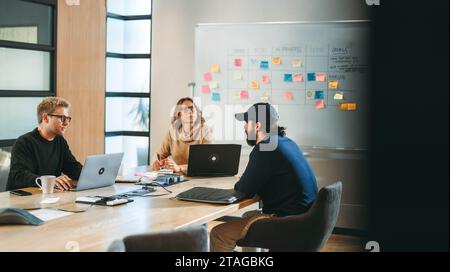 Professionisti aziendali eterogenei in un ufficio moderno si impegnano in una discussione collaborativa intorno a un tavolo da Consiglio di amministrazione. Utilizzo di notebook e puntamento verso una cornice Foto Stock