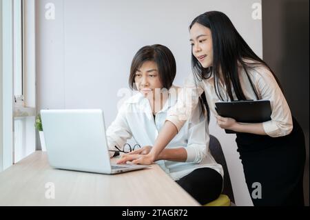 Una giovane donna d'affari asiatica sta aiutando una collega anziana a risolvere il problema del computer in ufficio. Foto Stock
