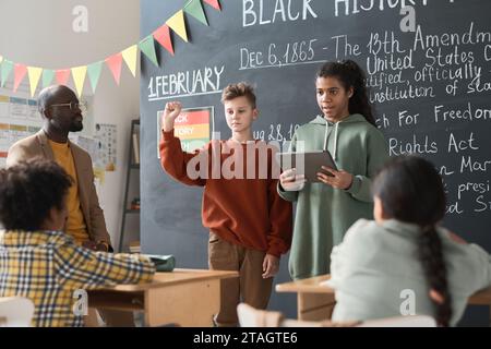 Due compagni di classe che utilizzano un tablet digitale per presentare il loro rapporto sul mese della storia nera durante una lezione mentre insegnanti e compagni di classe li ascoltano Foto Stock