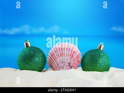 Conchiglia rosa con palline verdi lucenti natalizie sulla sabbia della spiaggia con il mare. Anno nuovo, vacanze. Copia spazio Foto Stock