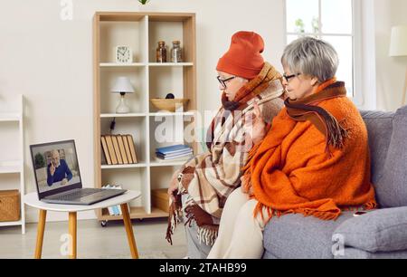 Coppia anziana ammalata o congelata seduta a casa con un computer portatile e con una videochiamata online con un uomo. Foto Stock