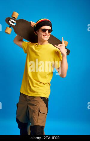 Ritratto di un ragazzo alla moda, vestito in stile reggae, rastaman e in posa sullo skateboard a spalla sullo sfondo blu dello studio. Foto Stock
