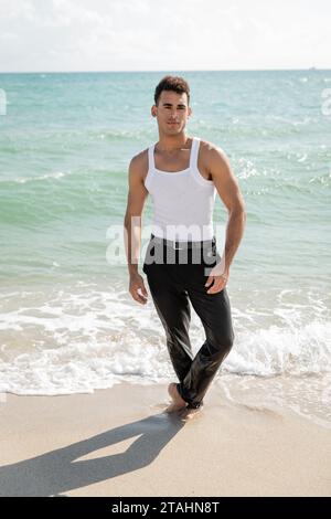 Uomo cubano che guarda la macchina fotografica mentre si trova sulla sabbia vicino all'acqua dell'oceano a Miami Foto Stock