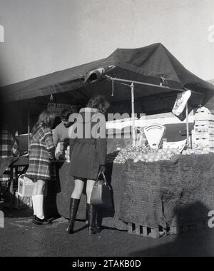 Anni '1970, storico, in un mercato agricolo, due acquirenti femminili in una bancarella all'aperto di frutta e verdura di J. W. Barfield & Son, Inghilterra, Regno Unito. I mercati degli agricoltori consentono ai coltivatori alimentari e alle imprese rurali locali di vendere i loro prodotti direttamente al pubblico. Foto Stock