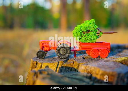 Un trattore giocattolo in miniatura si trova su un ceppo di alberi in un bosco, trasportando un piccolo albero nel suo letto. Foto Stock