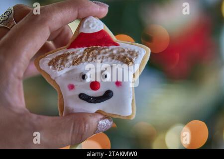 Mano di una donna che tiene un dolce biscotto a forma di stella di Natale decorato con un volto felice e un cappello di Babbo Natale. La messa a fuoco illumina lo sfondo Foto Stock