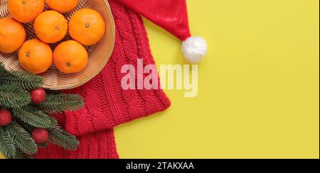 Mandarini in cesto, sciarpa in maglia rossa, cappello di Babbo Natale, rami di abete con palle di Natale su sfondo giallo. Composizione natalizia, anno nuovo, banne Foto Stock