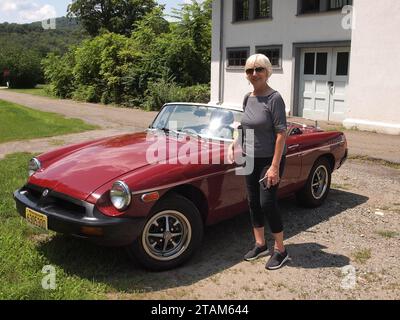 Donna di mezza età in piedi accanto a un roadster da 1977 MGB in Carmine Red. La capote è abbassata per godersi il sole. Foto Stock