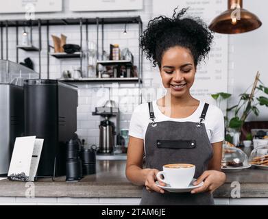 Barista sorridente in un grembiule che guarda una tazza di caffè. La donna con i capelli ricci lavora come barista in un bar. Foto Stock