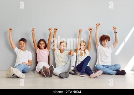 Gruppo di scolari seduti in fila sul pavimento su sfondo grigio con le mani al pugno. Foto Stock