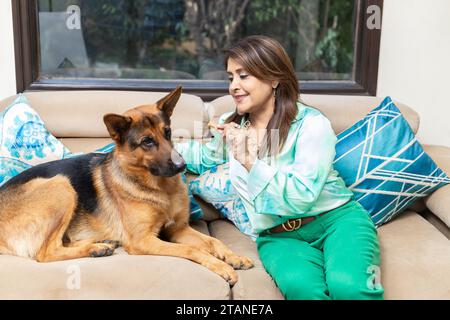 Bella donna indiana seduta su un divano che dà da mangiare al cane a casa. Foto Stock