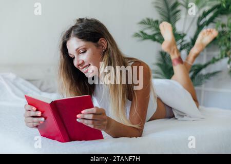 Bella donna snella in pigiama che legge un libro in camera da letto sul letto Foto Stock